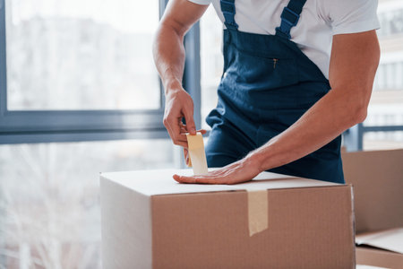 Packaging the box. Young male mover in blue uniform works indoors in the roomの写真素材