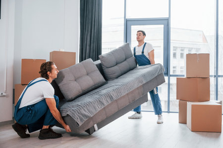 Carrying heavy sofa. Two young movers in blue uniform working indoors in the roomの写真素材