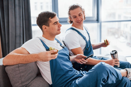 Sitting on the sofa. Tired and taking break. Two young movers in blue uniform working indoors in the roomの写真素材