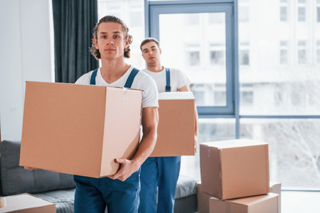 With boxes in hands. Two young movers in blue uniform working indoors in the roomの写真素材
