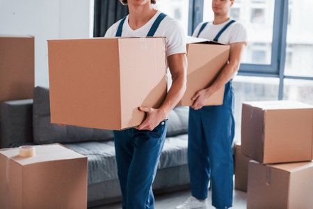 With boxes in hands. Two young movers in blue uniform working indoors in the roomの写真素材