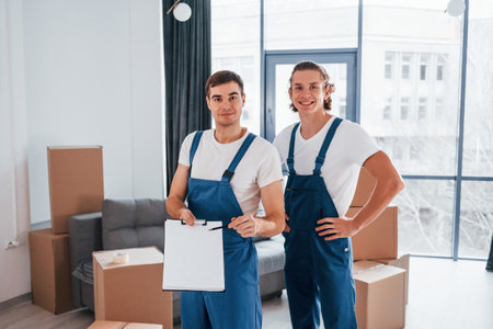 Holds notepad with document. Two young movers in blue uniform working indoors in the roomの写真素材