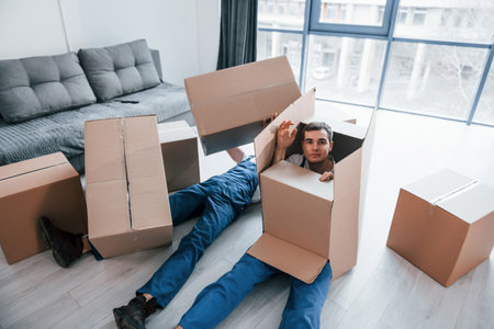 Two young male movers in blue uniform lying down on the floor with boxesの写真素材