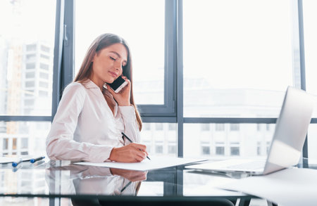 Have conversation by the phone. Young woman in white formal clothes is indoors in the modern officeの写真素材