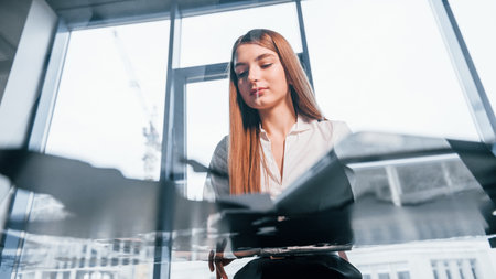 Sits by the table. Young woman in white formal clothes is indoors in the modern officeの写真素材