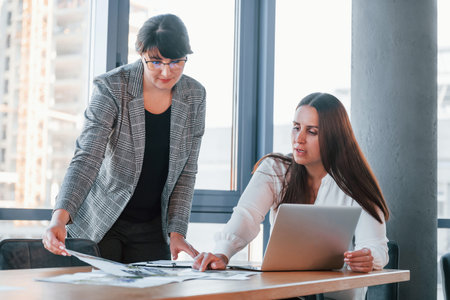 Using laptop. Two women in formal clothes is indoors in the modern office works togetherの写真素材