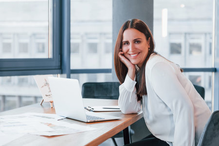 Sits by the table. Young woman in white formal clothes is indoors in the modern officeの写真素材