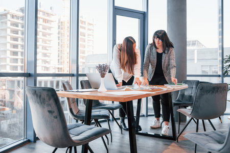 Standing and talking. Two women in formal clothes is indoors in the modern office works togetherの写真素材