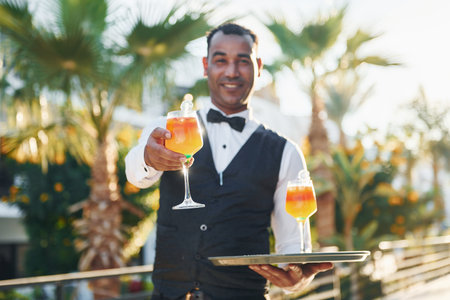 Holds cocktails. Black waiter in formal clothes is at his work outdoors at sunny daytimeの写真素材