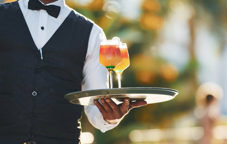 Holds cocktails. Black waiter in formal clothes is at his work outdoors at sunny daytimeの写真素材