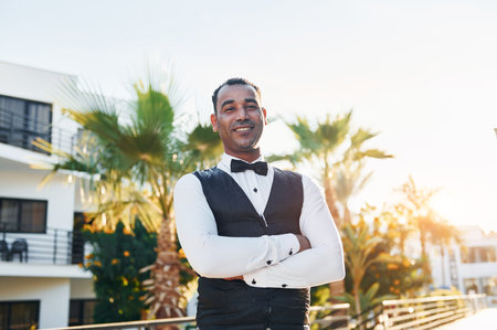 Posing for a camera. Black waiter in formal clothes is at his work outdoors at sunny daytimeの写真素材