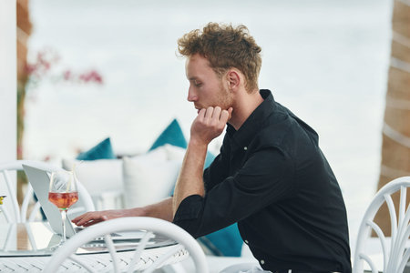 Sits by the table. Young man is outdoors at sunny daytime. Concept of vacation
