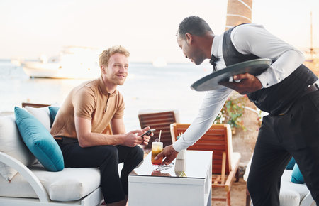 Serviced by waiter. Young man is outdoors at sunny daytime. Concept of vacation
