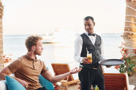 Serviced by waiter. Young man is outdoors at sunny daytime. Concept of vacation