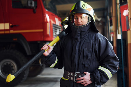 Holds axe in hands. Male firefighter in protective uniform standing near truckの写真素材