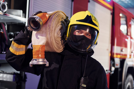Holds hose. Male firefighter in protective uniform standing near truck
