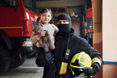 Holds teddy bear. Happy little girl is with male firefighter in protective uniformの写真素材