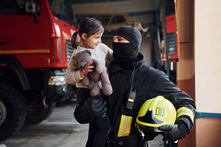 Holds teddy bear. Happy little girl is with male firefighter in protective uniformの写真素材