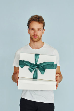 Young man in white shirt standing against wall and holds gift boxの写真素材
