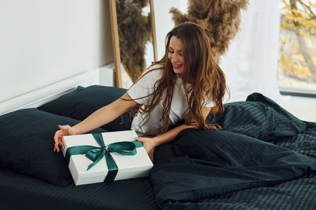 Gift box in hands of young woman. Interior and design of beautiful modern bedroom at daytimeの写真素材