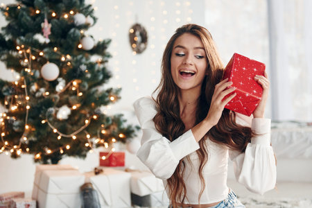 Happy young woman sits indoors near christmas tree and celebrates New yearの写真素材