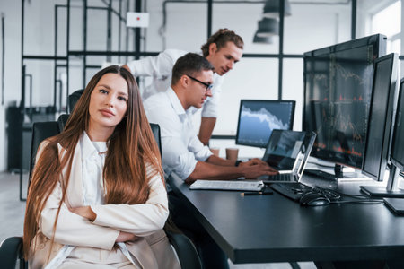 Woman looks into the camera. Team of stockbrokers works in modern office with many display screens