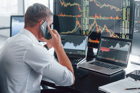 Talks by phone. Young male stock brocker works indoors in the office. Many of displaysの写真素材