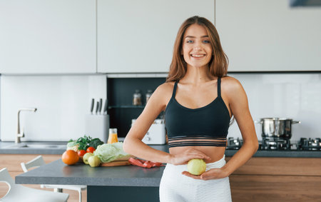 Holds apple against belly. Young european woman is indoors at kitchen indoors with healthy foodの写真素材