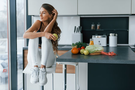 Sits and holds apple. Young european woman is indoors at kitchen indoors with healthy foodの写真素材
