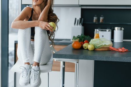 Sits and holds apple. Young european woman is indoors at kitchen indoors with healthy foodの写真素材