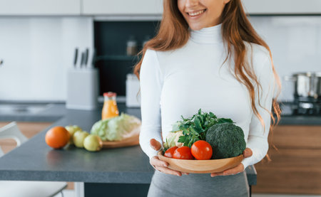 Holds plate with vegetables. Young european woman is indoors at kitchen indoors with healthy foodの写真素材