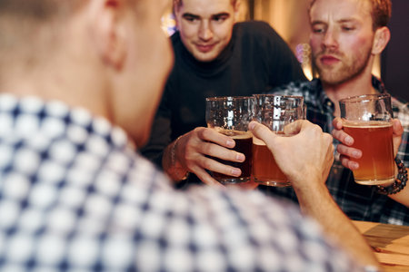 Cheering by knocking beer glasses. Group of people together indoors in the pub have fun at weekend timeの写真素材