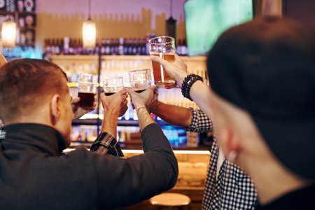Cheering by knocking beer glasses. Group of people together indoors in the pub have fun at weekend timeの写真素材