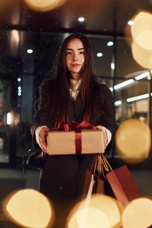 Standing near building and holds gift box. Cheerful woman is outdoors at Christmas holidays time. Conception of new yearの写真素材