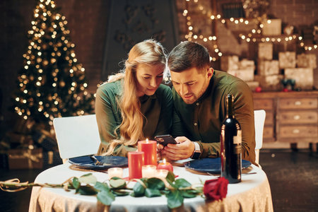 Sits and using phone. Young lovely couple have romantic dinner indoors togetherの写真素材