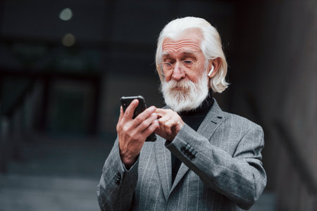 Senior businessman in formal clothes, with grey hair and beard is outdoors trying wireless headphones with phoneの写真素材
