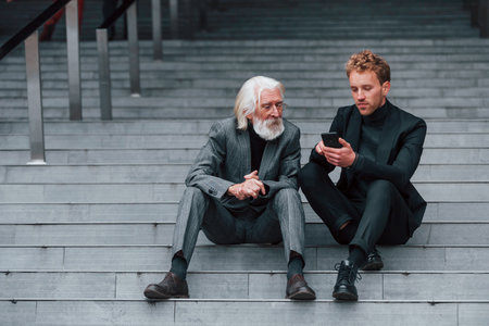 Sitting with phone. Young guy with senior man in elegant clothes is outdoors together. Conception of businessの写真素材