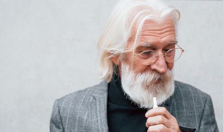 Senior businessman in formal clothes, with grey hair and beard is outdoors smokes electronic cigaretteの写真素材