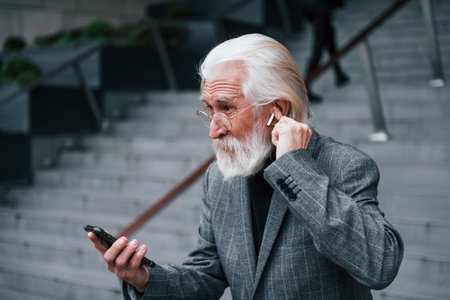 Senior businessman in formal clothes, with grey hair and beard is outdoors trying wireless headphones with phoneの写真素材