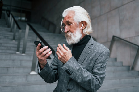 Using smartphone. Senior businessman in formal clothes, with grey hair and beard is outdoorsの写真素材