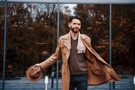 Standing against building with glass doors. Young male model in fashionable clothes is outdoors in the city at daytimeの写真素材