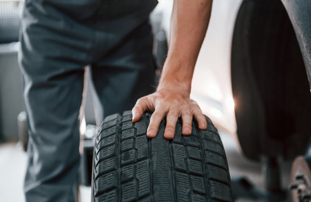 Works with tire. Adult man in grey colored uniform is in the automobile salonの写真素材