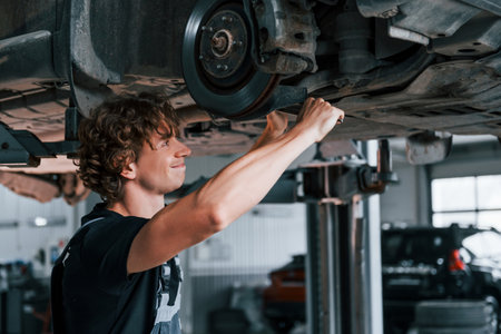 Uses wrench to fix car. Adult man in grey colored uniform works in the automobile salonの写真素材