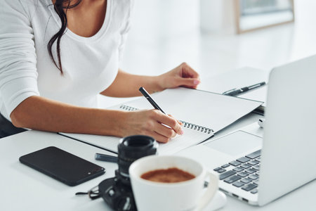 Using laptop and notepad. Young female freelancer working indoors in the office at daytimeの写真素材