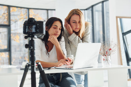 Camera on tripod is recording. Two young female freelancers working indoors in the office with laptopの写真素材