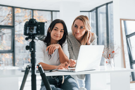 Camera on tripod is recording. Two young female freelancers working indoors in the office with laptopの写真素材