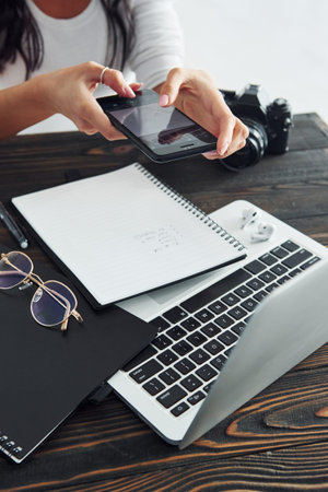 Holding phone. Young female freelancer working indoors in the office at daytime. Laptop and notepad