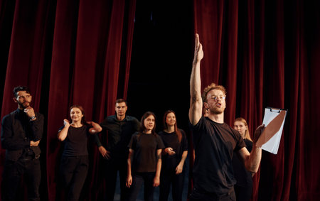 Man with notepad practice his role. Group of actors in dark colored clothes on rehearsal in the theaterの写真素材