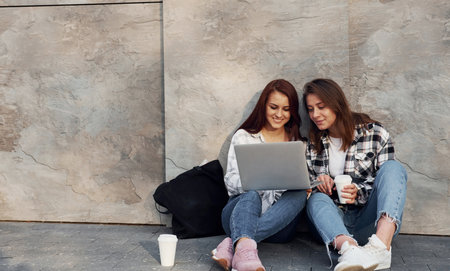Using laptop. Beautiful cheerful friends or lesbian couple together near wall outdoors at daytimeの写真素材
