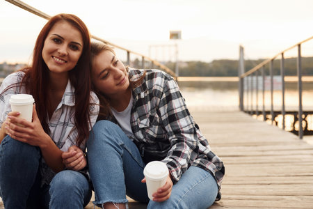 Cute lesbian couple sits together near the lake with cups of drink in hands and embracing each otherの写真素材